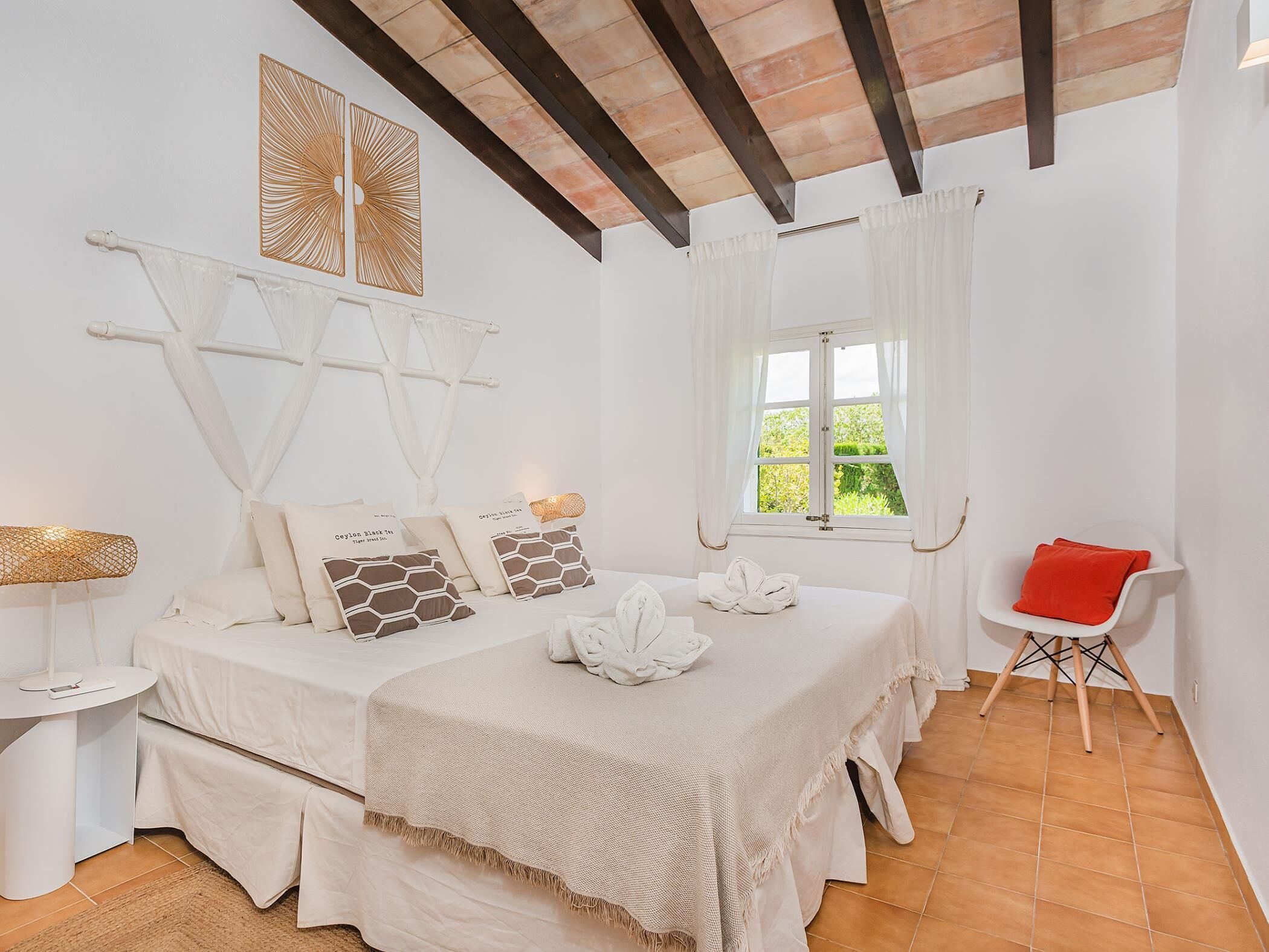 Ein Schlafzimmer mit Bett, Nachttisch, Sessel, Fenster mit Vorhängen und Decke mit sichtbaren Balken.