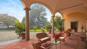 Un patio techado con mobiliario de mimbre, plantas en macetas y vista a un jardín.