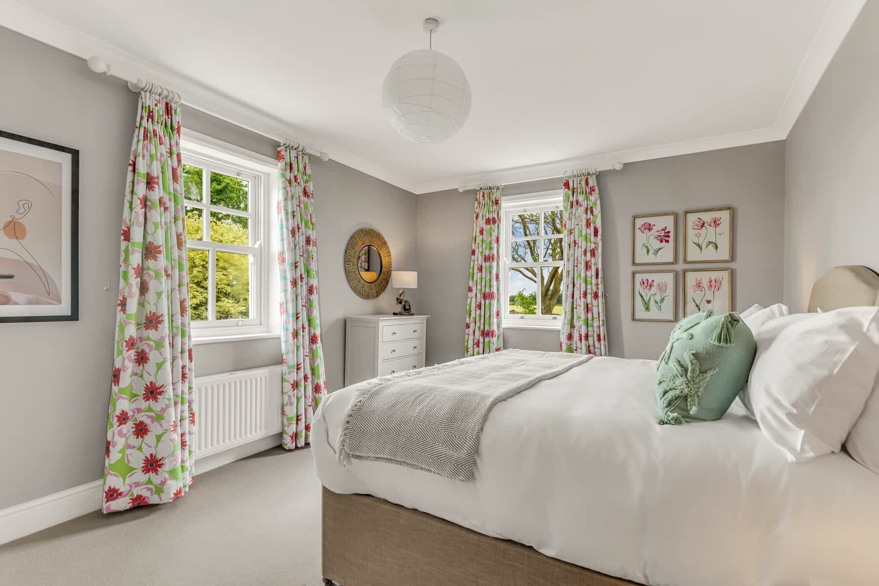 Een slaapkamer met een bed, twee ramen met bloemen gordijnen, een nachtkastje met een lamp, en bloemistisch kunstwerk in een lijst aan de muur.