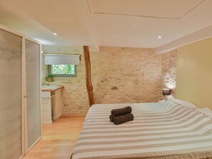 A bedroom with a bed, a wooden floor, a stone wall, and a window with a view of greenery.