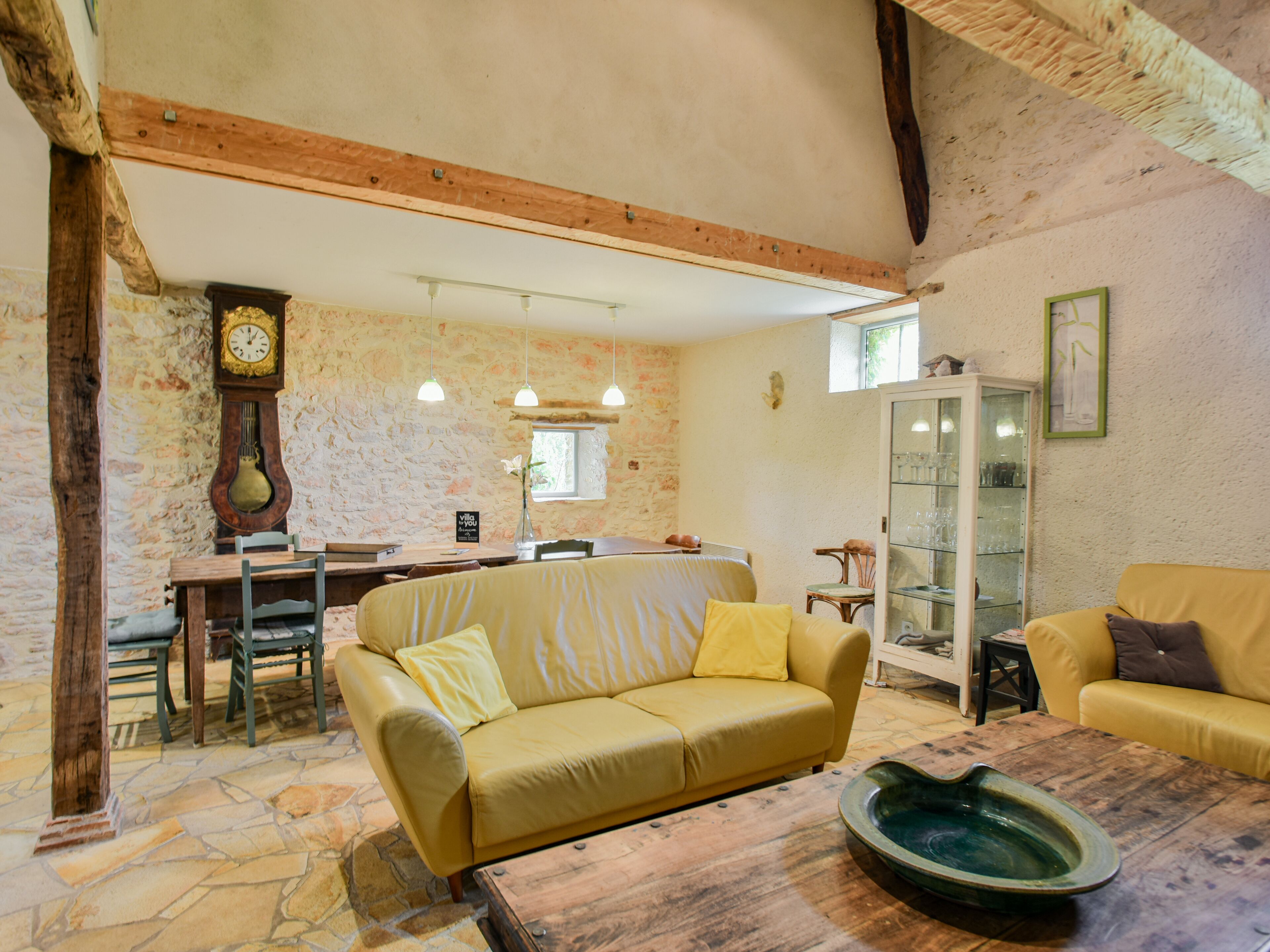 A cozy living room with a yellow sofa, a wooden coffee table, and a stone floor. There is a dining area with a wooden table and chairs, and a kitchen visible in the background.