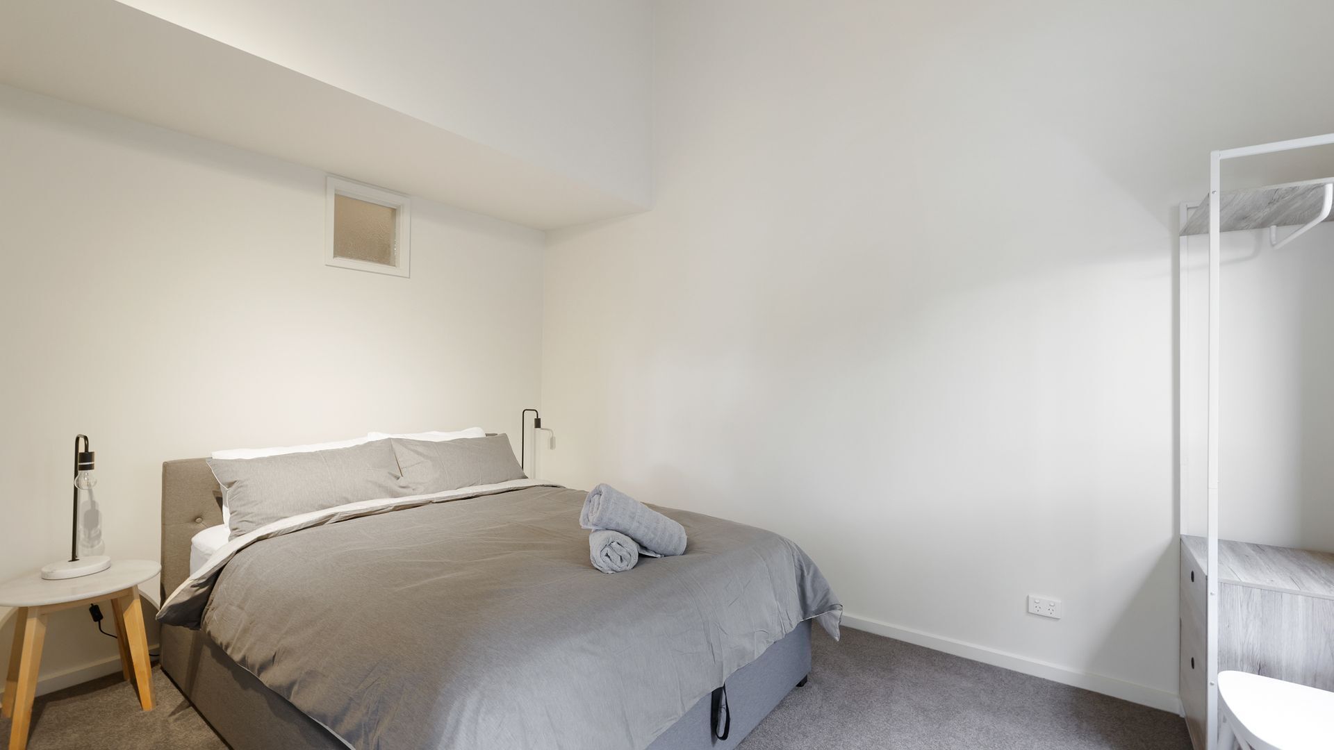 Une chambre à coucher comprenant un lit, une table de chevet, une lampe, une prise de courant et une petite fenêtre.