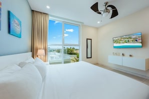 A hotel room with a large bed, a TV, a ceiling fan, and a balcony with a view of the beach.