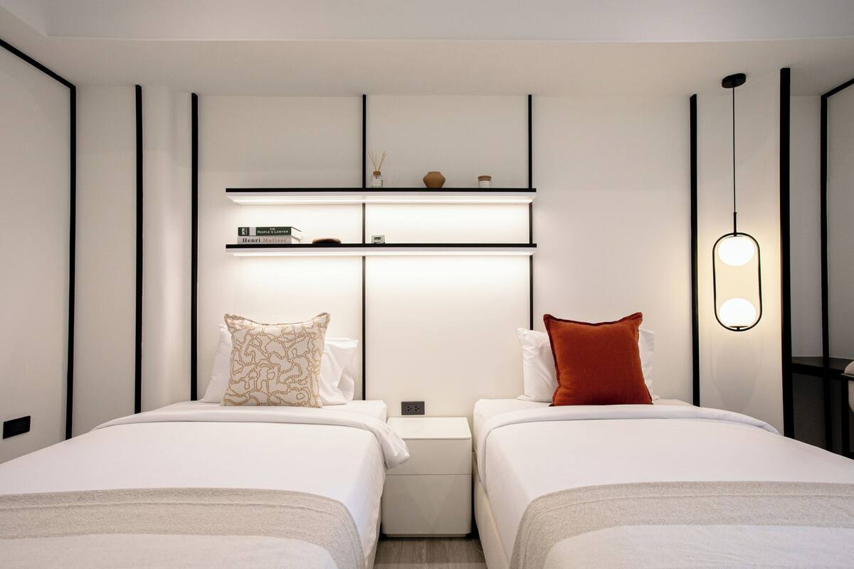 A modern hotel room with two beds, a built-in shelf with decorative items, a bedside table, and a wall-mounted light fixture.