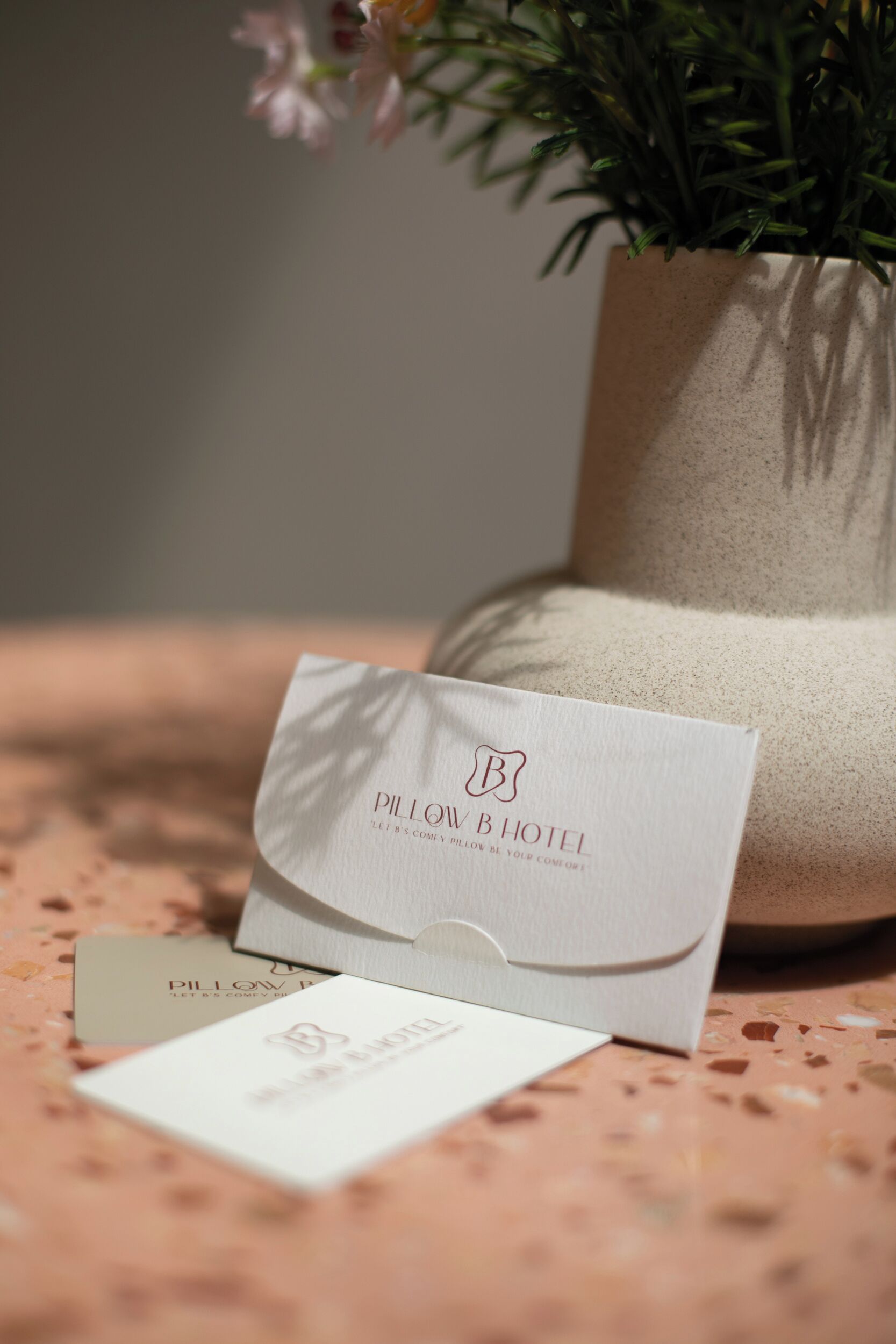 A white business card with the text "Pillow & Hotel" and a logo, placed on a speckled surface with a potted plant in the background.