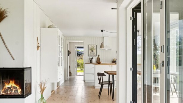 A modern kitchen with white cabinets, a dining area with a wooden table and chairs, and a fireplace with a visible fire. - "Ødke" - 350m from the sea by Interhome (Odder)