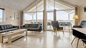 A spacious living area with a wooden floor, a sofa, a chair, and a dining table. Large windows offer a view of the outdoors. - Amazing home in Jerup with sauna (Jerup)