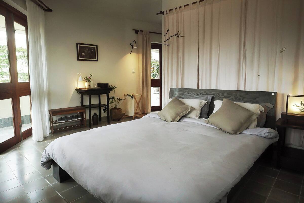A bedroom with a large bed, a bedside table, a chair, and a window with curtains.
