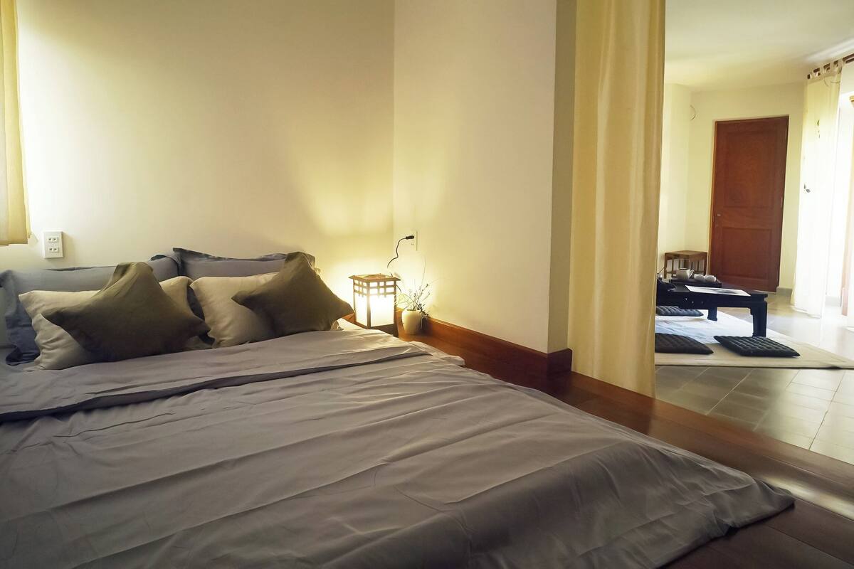 A hotel room with a large bed, a nightstand with a lamp, and a view of the hallway.