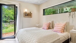 Une chambre avec un grand lit, une table de chevet, une étagère fixée au mur et une fenêtre donnant sur un paysage verdoyant.