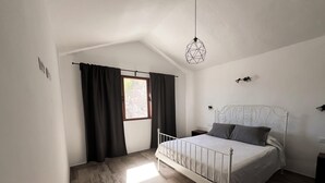 A bedroom with a bed, a window with curtains, a hanging light fixture, and a wooden floor.