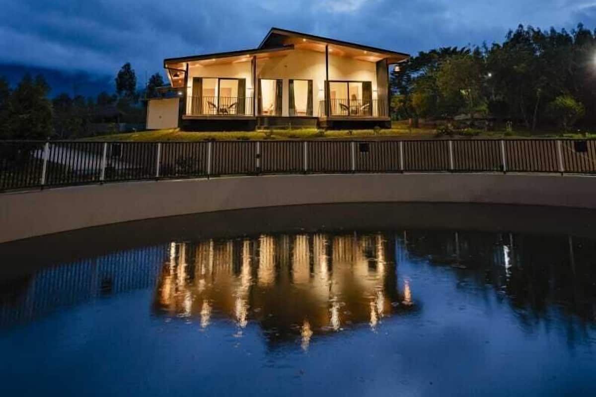 A modern house with a large balcony, a swimming pool in the foreground, and a cloudy sky in the background.