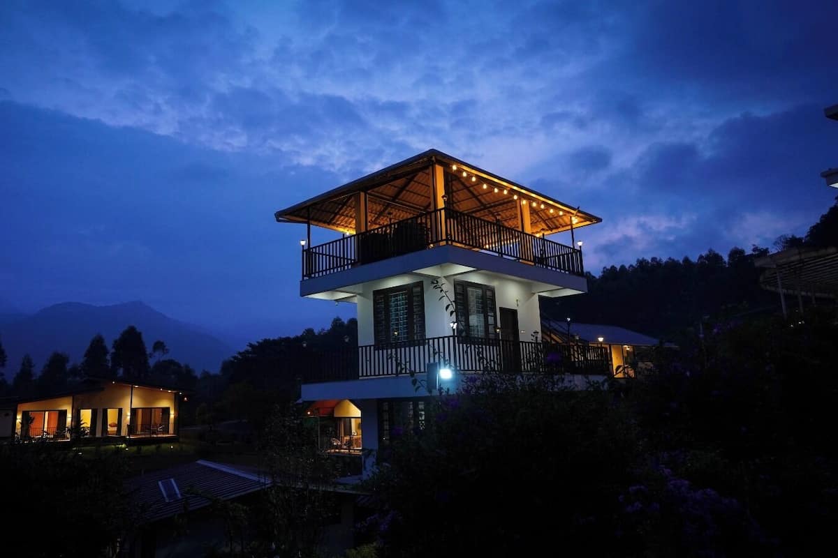 A multi-story building with a balcony on the upper floor, surrounded by trees and mountains in the background.