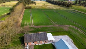 Oversigtsbillede af en landejendom med et hus, grønne marker og et vandområde i baggrunden.