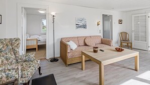 En rummelig stue med en stor sofa, et sofabord og en fletstol. I det tilstødende værelse er der en seng, en gulvlampe og et billede på væggen.