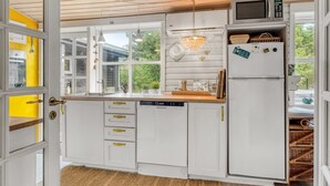 Moderni keittiö, jossa on valkoiset kaapit, mikroaaltouuni ja jääkaappi. Suuri ikkuna tuo luonnonvaloa ja avautuu vehreään maisemaan.