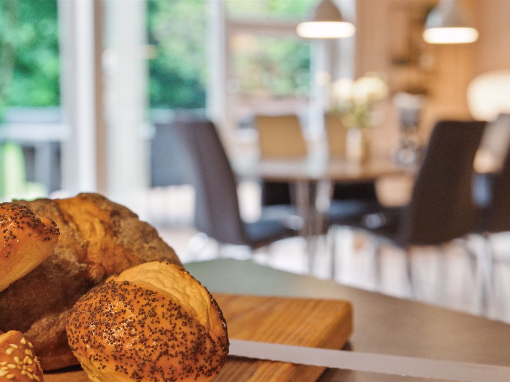Un croissant su un tagliere di legno con un coltello, in un ambiente interno ben illuminato con una sala da pranzo sullo sfondo.