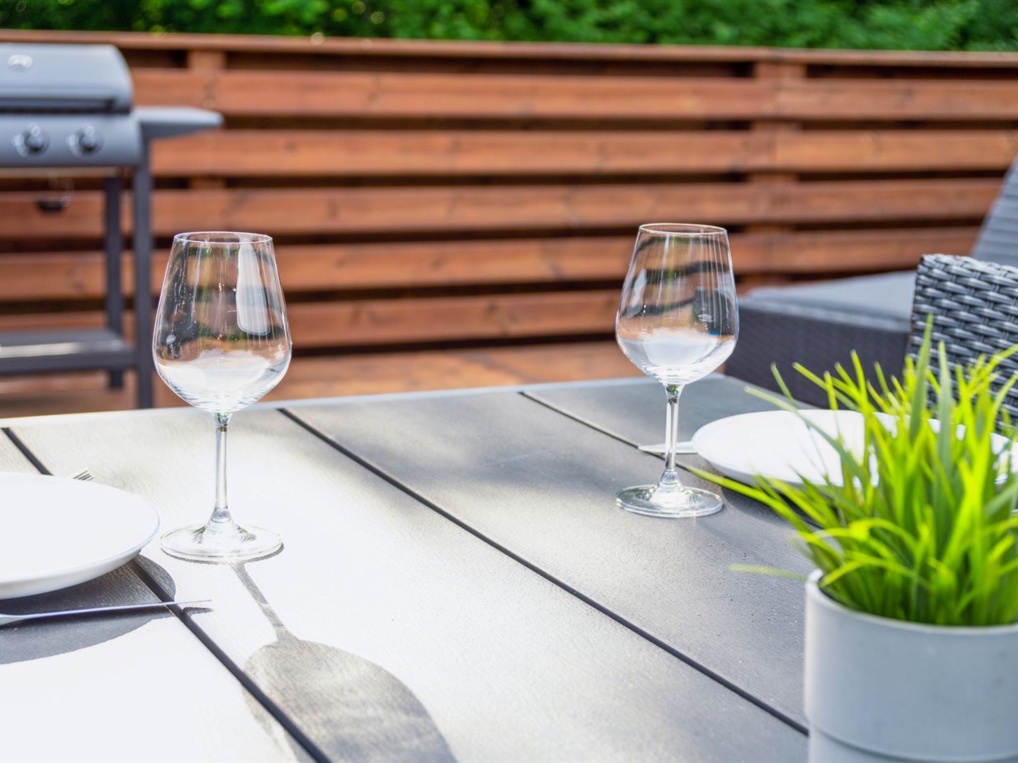 An outdoor dining area with two wine glasses on a table, a potted plant, and a grill in the background.