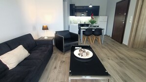 A modern living room with a black sofa, a small side table with a lamp, and a coffee table with decorative items. A kitchen is visible in the background with a dining area. - Highlighted Retreat (Athens)