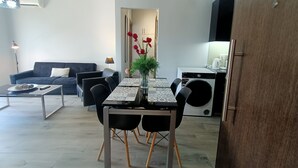 A modern living room with a dark blue sofa, a dining table with chairs, a coffee table, and a small kitchen area with a washing machine. - Highlighted Retreat (Athens)