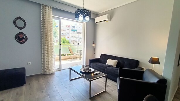 A modern living room with a sofa, a coffee table, and a chair. There is a balcony with a view of buildings and greenery. - Highlighted Retreat (Athens)