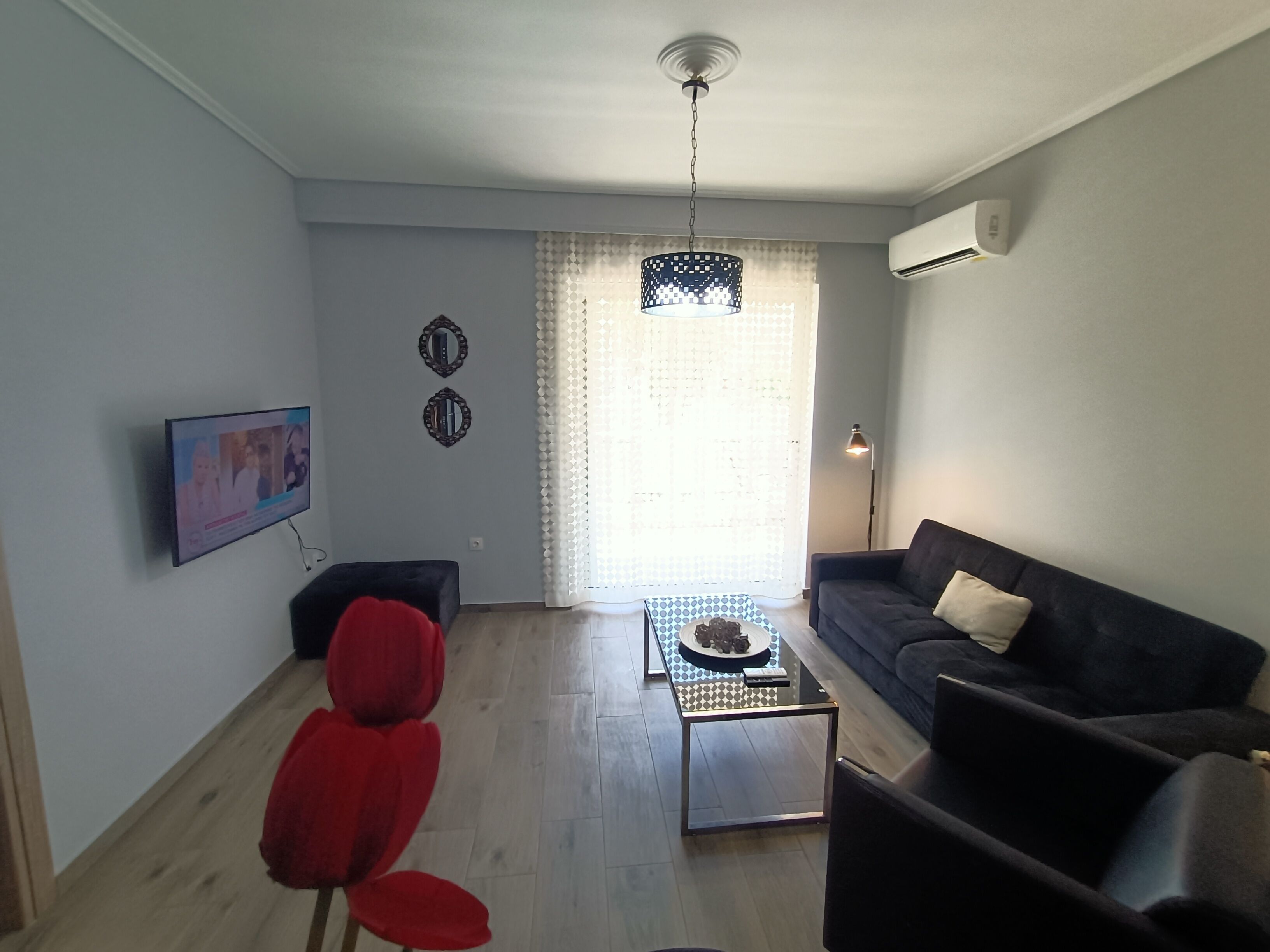 A living room with a sofa, a red chair, a coffee table, a TV mounted on the wall, and a window with curtains.