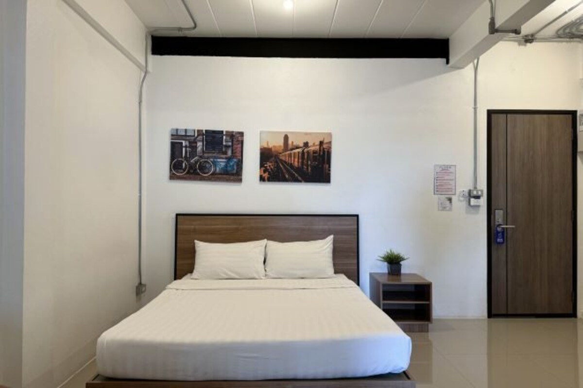 A hotel room with a bed, two wall-mounted artworks, a small bedside table with a plant, and a door.
