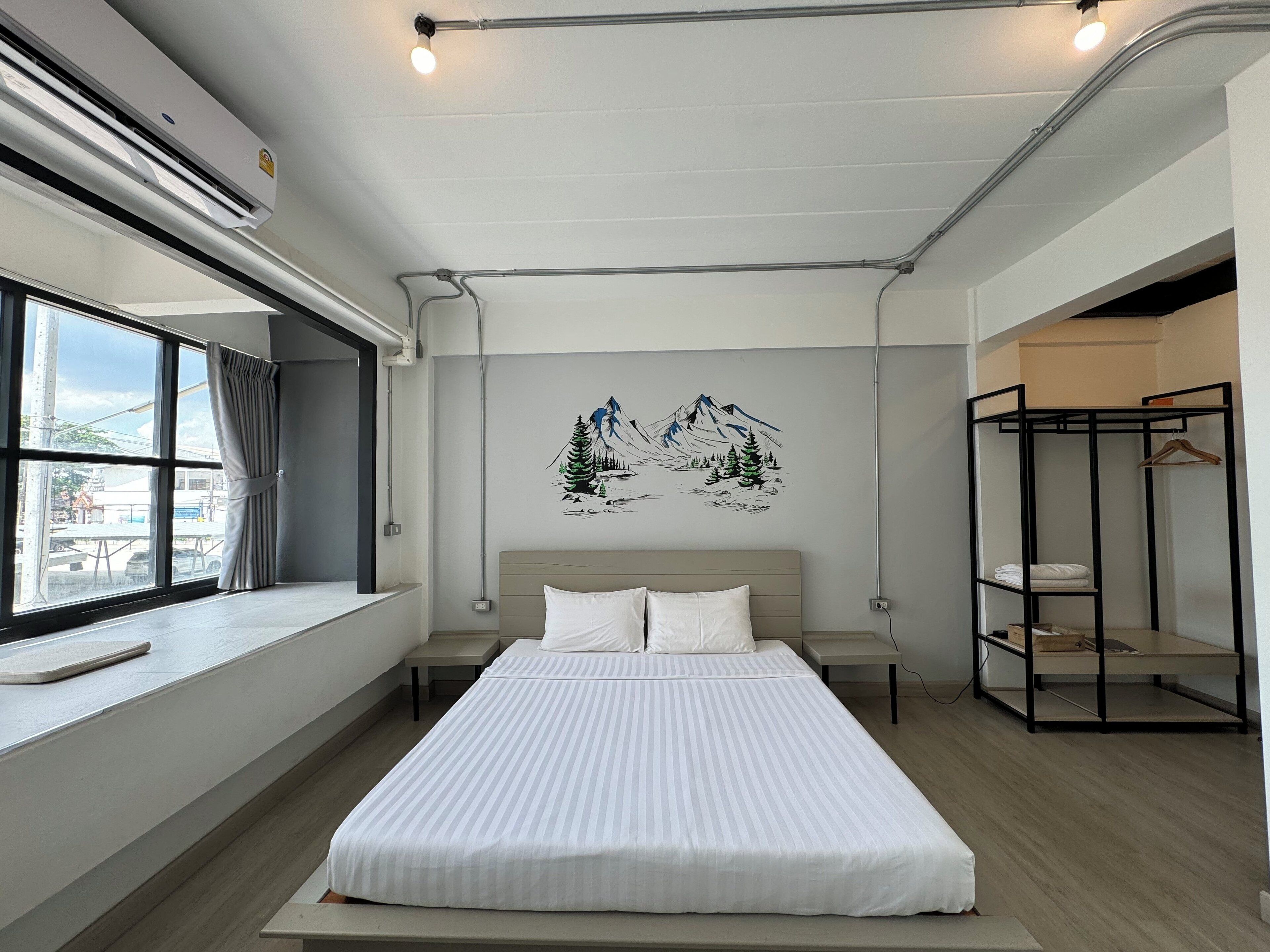 Una habitación de hotel moderna con una cama grande, una ventana con cortinas, un cuadro de montañas colgado en la pared y un armario metálico.