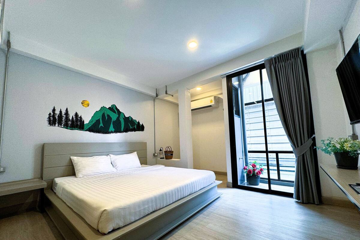 A modern hotel room with a large bed, a mountain mural on the wall, a window with curtains, and a potted plant on the windowsill.
