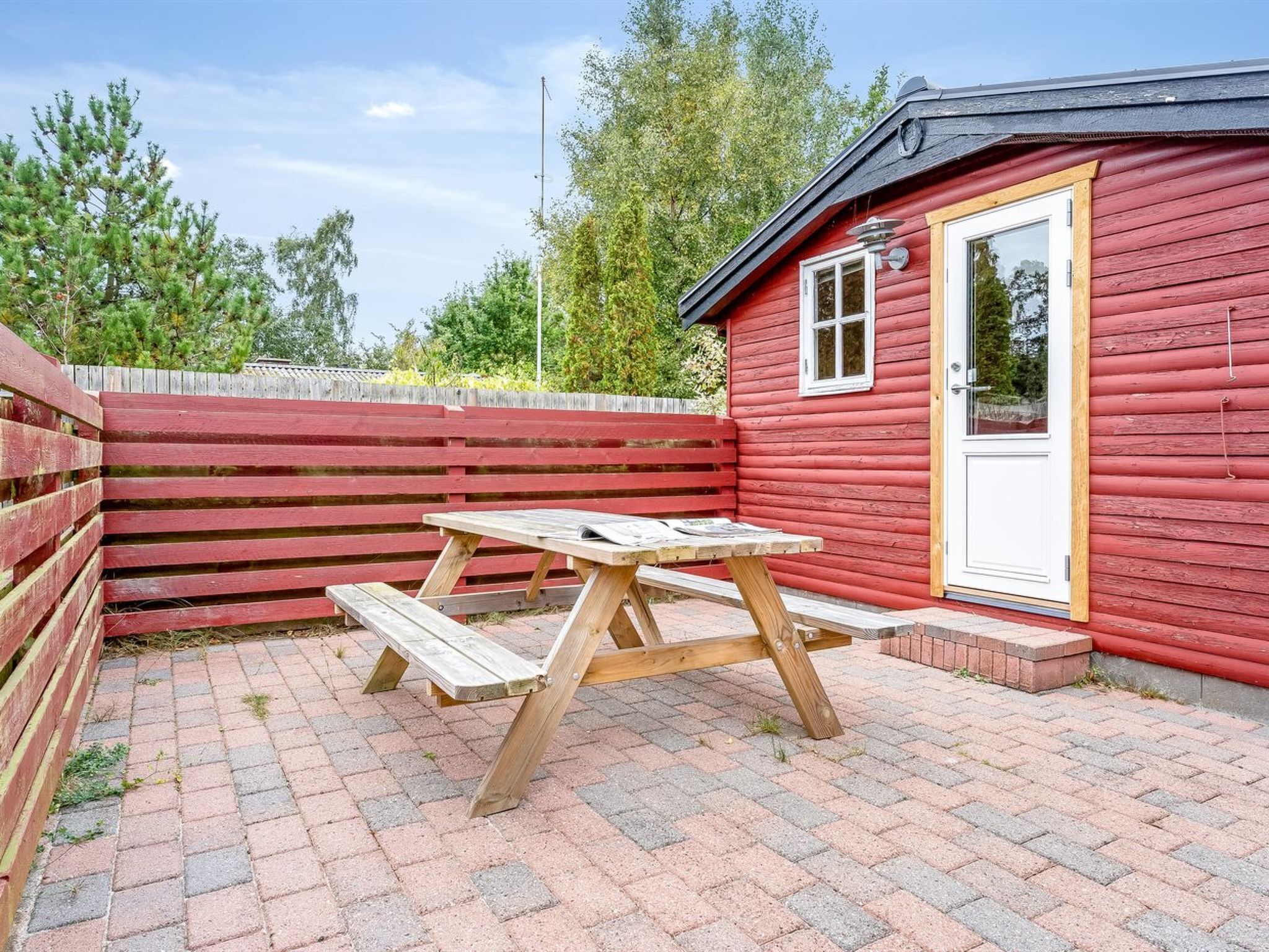 Une cabane en bois rouge, avec une table de pique-nique et des bancs, entourée d’une clôture en bois et d’un chemin pavé de briques.