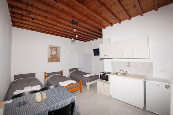 A small, clean room with two beds, a wooden ceiling, a kitchenette, and a table with a blue tablecloth. - Fournakia Studio (Mykonos)