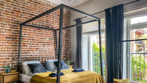 A four-poster bed with a mustard-colored bedspread, a wooden nightstand with a vase, and a view of a brick wall and greenery outside.
