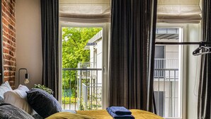 A bedroom with a large window, a bed with a mustard-colored bedspread, and a view of a balcony and greenery outside.