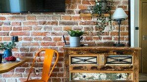 A room with a brick wall, a flat-screen TV mounted above a wooden cabinet, a chair, and a table with a plant and a bowl of fruit.