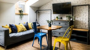 A modern living room with a loft-style ceiling, a grey sofa with yellow and blue cushions, a wooden dining table, and a TV mounted on a brick wall. - Cozy Apartment with A/C for couples, City Center 2, Różana (Kraków)