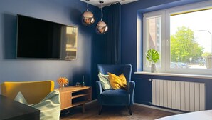 Un salon moderne avec un mur bleu, un téléviseur fixé au mur, un fauteuil bleu, une table basse en bois et une fenêtre donnant sur la rue.