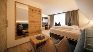 A modern hotel room with a large bed, a sitting area with a sofa and a small table, a TV mounted on a wooden wall, and a large mirror.