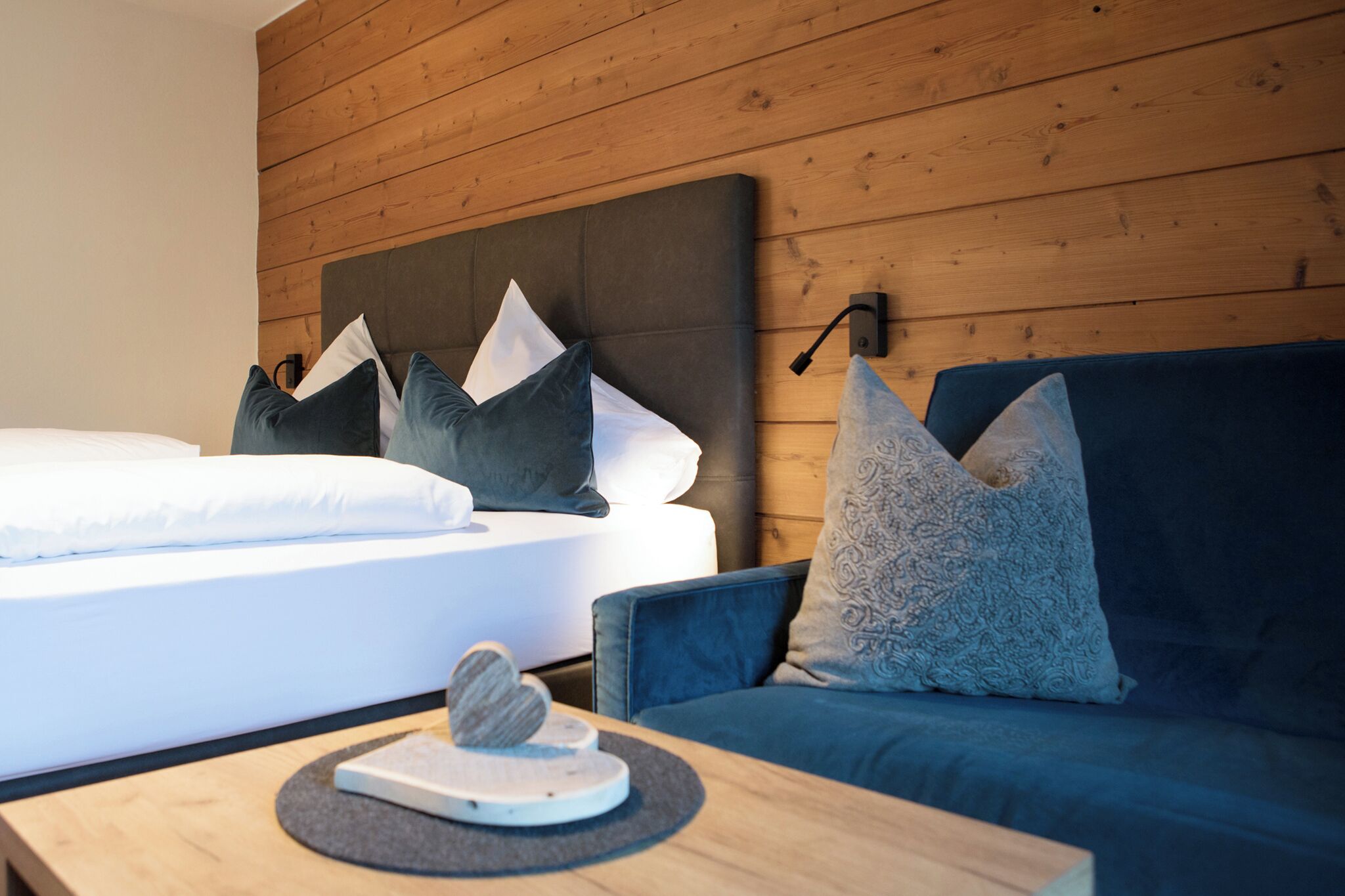 Une chambre d’hôtel moderne avec un panneau mural en bois, un lit avec du linge de lit blanc et des oreillers foncés, un canapé bleu avec des coussins à motifs et une petite table en bois sur laquelle est posé un objet décoratif.