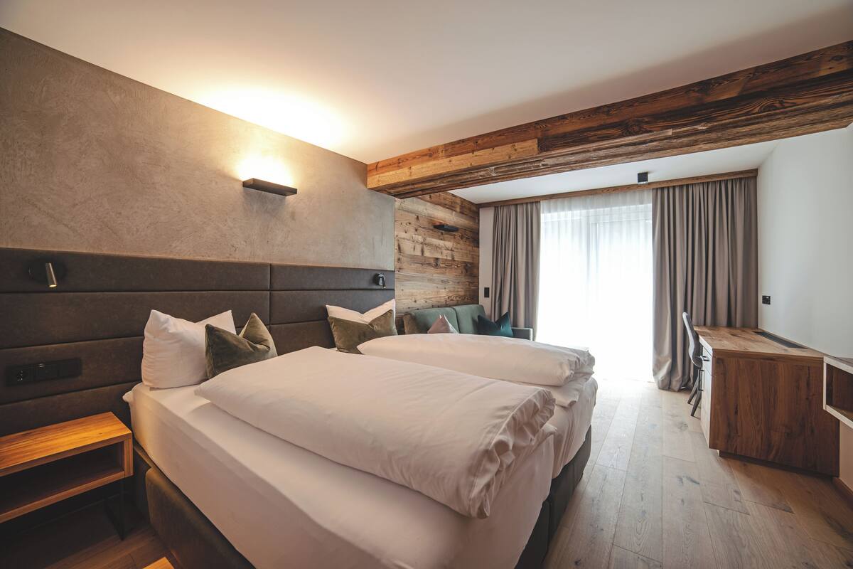 A modern hotel room with two beds, a wooden headboard, a built-in desk, and a large window with curtains.