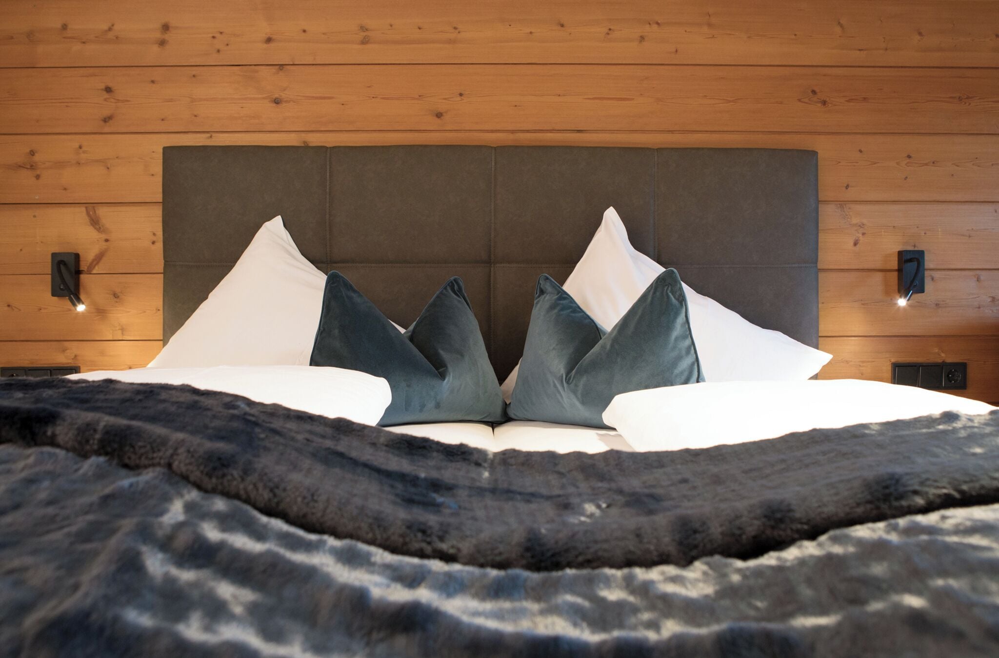 Una cama con cabecera oscura, almohadas blancas y oscuras, y una manta oscura con textura.