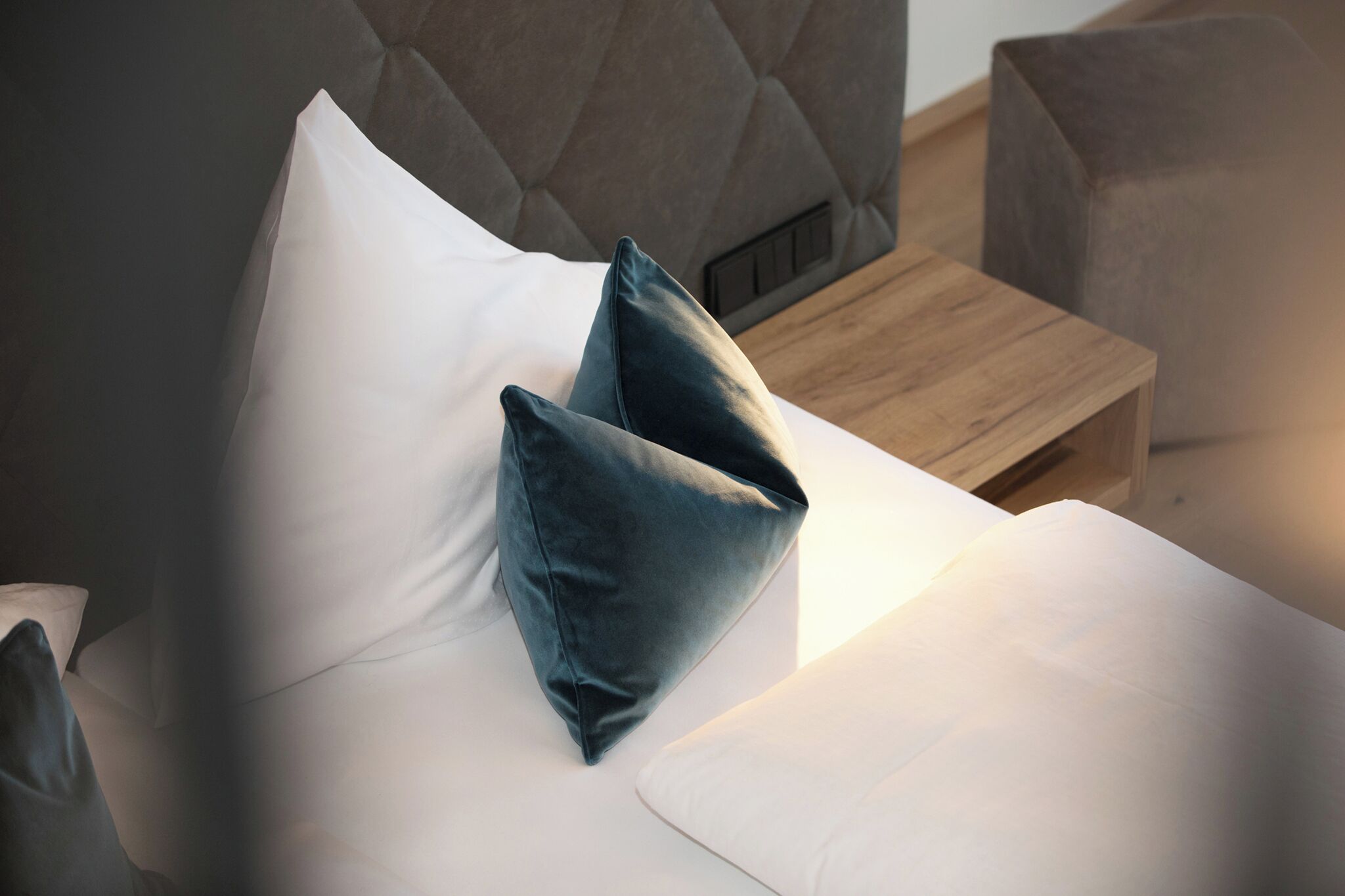 Un lit avec du linge de lit blanc et deux oreillers foncés, une table de chevet en bois et une tête de lit capitonnée.
