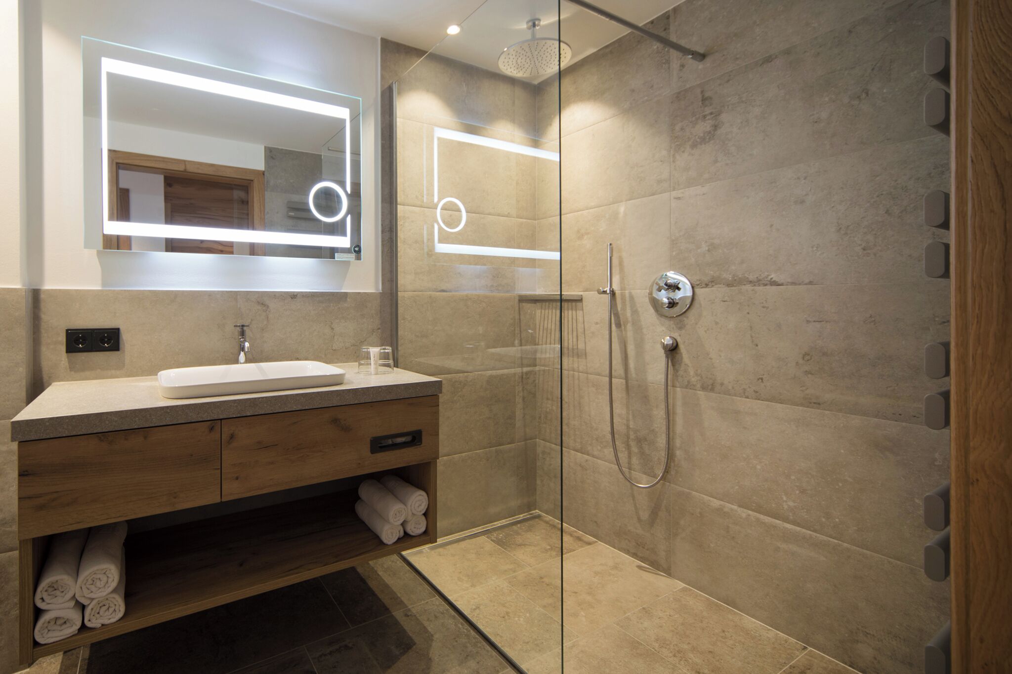 A modern bathroom with a glass-enclosed shower, a wooden vanity with a mirror, and a built-in shelf for towels.