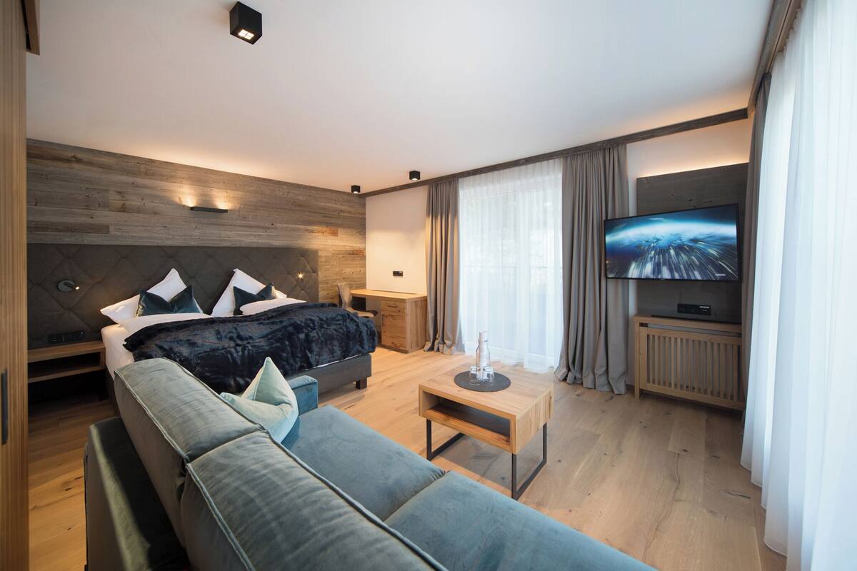 A modern hotel room with a large bed, a sofa, a TV, and a wooden floor.