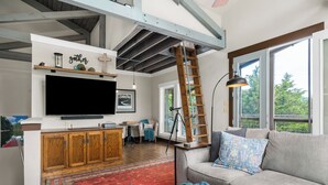 Een ruime woonkamer met een grote flatscreen-televisie, houten kasten, een vloerkleed met een rood patroon en een grijze bank met decoratieve kussens.