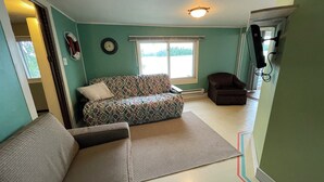 Un salon avec un canapé à motifs, un petit divan et un fauteuil. On y trouve une fenêtre avec des stores, une horloge murale et un ventilateur de plafond.