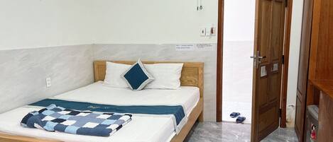 A single-bed room with a wooden headboard, a bedspread, and a blue and white checkered blanket. There is a wooden door to the right and a small wooden cabinet below it.
