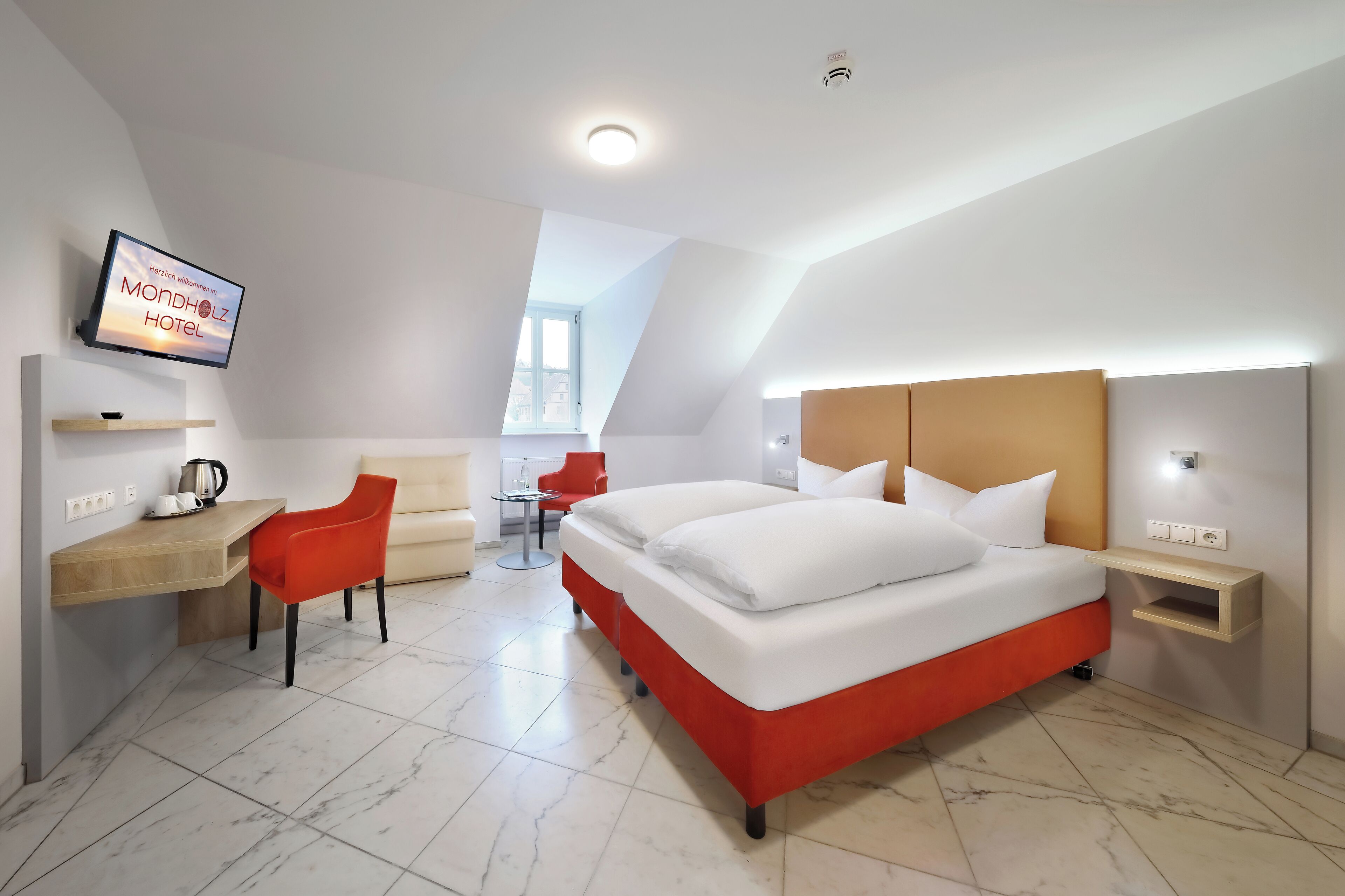 A modern hotel room with a large bed, a desk, a chair, and a television displaying "Mondholz Hotel".