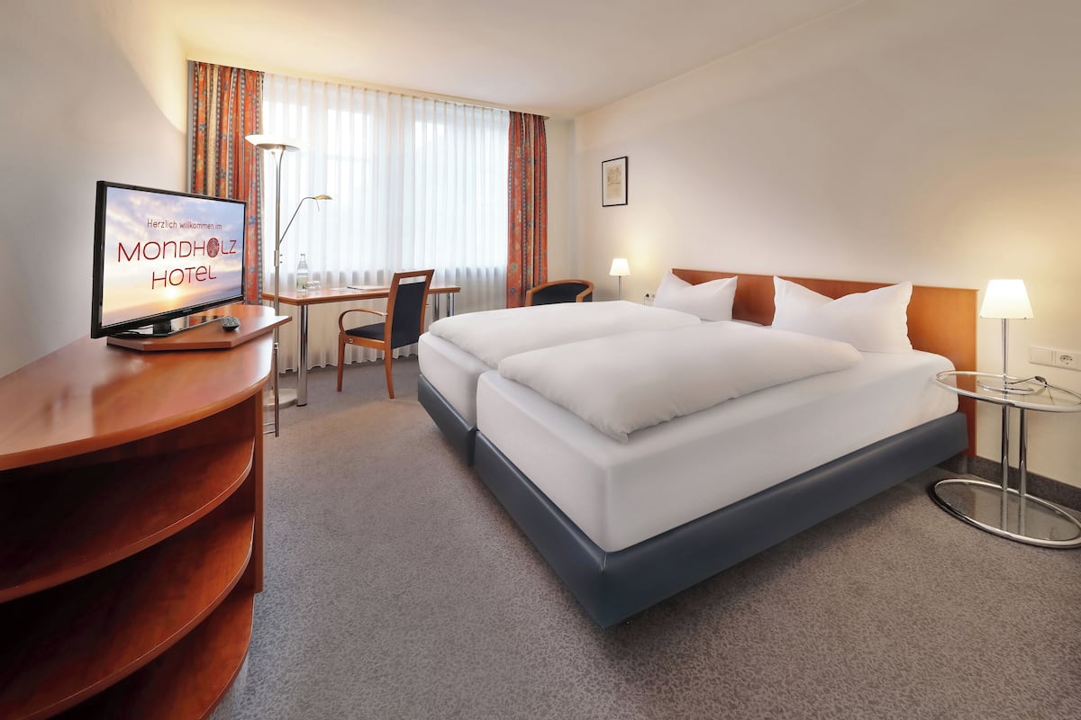 A hotel room with a bed, a desk with a computer, a chair, a lamp, and a window with curtains.