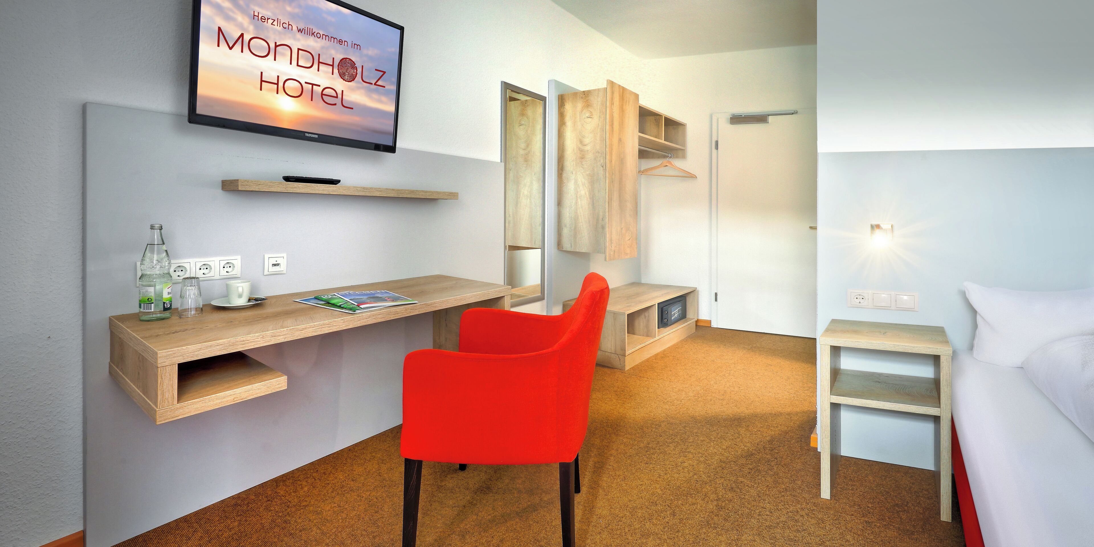 Et hotelværelse med et fjernsyn, der viser 'Herzlich willkommen im MONHOLZ HOTEL', et træskrivebord, en rød stol, et natbord og en seng.