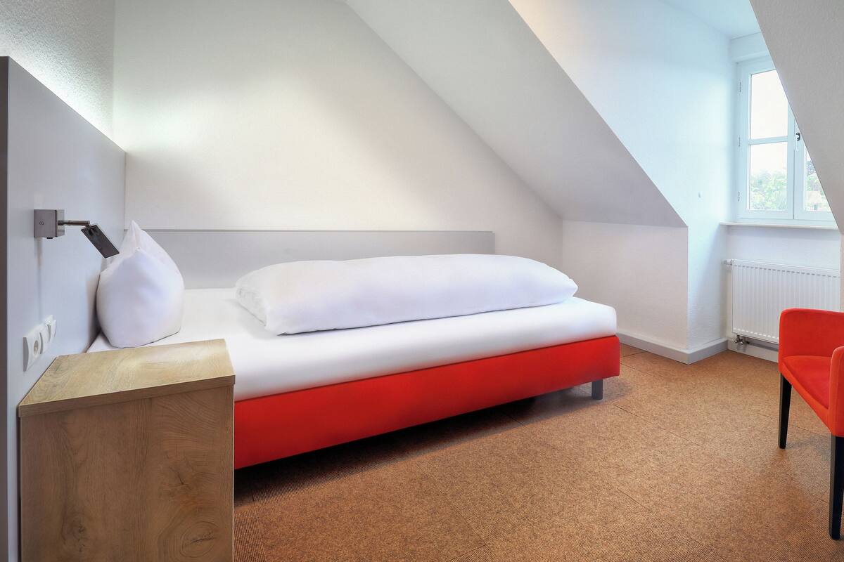 A modern room with a bed, a red chair, a wooden side table, and a window.